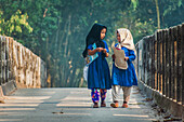 Bogura, Bangladesh - 24 November 2018: View of two young girls in vibrant blue dresses and contrasting headscarves walk on a bridge, sunlight dappling the scene.