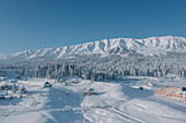 Aerial view of snow-laden trees and buildings blanketed in white against a backdrop of majestic, snow-capped mountains, Gulmarg, Jammu and Kashmir, India.