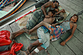 Dhaka, Bangladesh - 15 August 2021: View of men and children resting on a wooden platform near boats, their exhausted forms a stark contrast to the vibrant colors of their clothing.