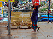 Dhaka, Bangladesh - 17 May 2019: View of a woman carrying a heavy box overhead, walking along the Buriganga River's edge, amidst the vibrant chaos of boats and riverside dwellings.