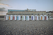 View of colorful beach huts stand in a neat row against the backdrop of a grand building under a pale sky, a tranquil scene of coastal charm, Le Tréport, Normandy, France.