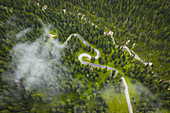 Aerial view of winding roads embraced by a sea of emerald green trees, with low-lying clouds adding a mystical touch to the landscape, Cortina d'Ampezzo, Veneto, Italy.