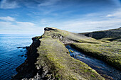 Blick auf zerklüftete Klippen und einen schmalen Bach, der unter einem klaren Himmel auf den weiten, tiefblauen Ozean trifft und eine ruhige und zugleich dramatische Landschaft schafft, Ketubjörg, Island.