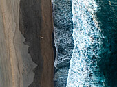 Aerial view of dark sands meeting the turbulent, frothy blues of the Atlantic waters, creating a stark contrast of textures and hues, Black Beach, Azores, Portugal.