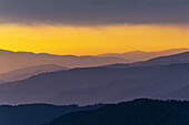 Blick auf geschichtete Bergsilhouetten in Blau- und Violetttönen vor einem feurigen orange-gelben Himmel in der Abenddämmerung, Steiermark, Österreich.