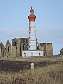 View of the striking Phare de Saint-Mathieu lighthouse rises boldly amidst ancient ruins, a solitary figure wanders through golden fields, Plougonvelin, Bretagne, France.