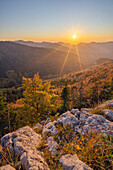View of the sun bursting over the forested, mountainous landscape, where autumn paints the trees in warm hues of orange and gold, Blatnica, Žilina Region, Slovakia.
