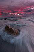 View of a solitary rock enduring the relentless caress of foamy waves beneath a dramatic, crimson-streaked sky, a tranquil dance of color and motion, Cape Town, Western Cape, South Africa.