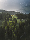 Aerial view of a solitary cabin glows amidst the verdant forest, nestled in a meadow kissed by the morning mist, Alpe di Siusi, Trentino, Italy.