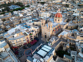 Aerial view of the vibrant square with its bustling activity and the imposing church with its striking red dome, against the backdrop of the dense urban landscape, Gozo, Malta.