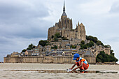 Mont Saint-Michel, Frankreich - 19. August 2025: Blick auf ein Kind, das im nassen Sand spielt, während sich die mittelalterliche Abtei majestätisch gegen den bewölkten Himmel abhebt.