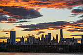 Blick auf die Silhouette der Skyline von Manhattan vor einem feurigen Sonnenuntergang mit orange und violett leuchtenden Wolken, die sich im ruhigen Wasser spiegeln, New York, New York, Vereinigte Staaten.