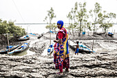 Shyamnagar, Bangladesch - 13. Mai 2023: Blick auf eine Frau in farbenfroher Kleidung, die auf schlammigem Gelände in der Nähe von geparkten Booten steht, mit einem Kind, das am Flussufer spielt, alles unter einem bewölkten Himmel.