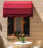 Bagno Vignoni, Tuscany, Italy - 28 April 2025: View of a quaint window adorned with a vibrant red awning, casting shadows on a table set for two, a scene of understated elegance.