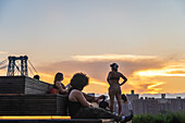 Domino Park, Brooklyn, United States - 12 August 2025: View of golden light painting the sky above the Williamsburg Bridge as people relax on wooden tiers, creating a tranquil urban scene.