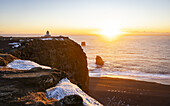View of the sun kisses the dark beach and rugged cliffs where a lighthouse stands guard in the golden light, Vik, Mýrdalshreppur, Iceland.