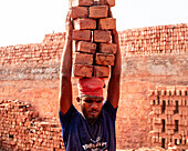 Narsingdi, Bangladesh - 08 December 2024: View of a worker with a red cloth tied on his head carrying a stack of bricks against the backdrop of a brick kiln.