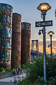 Domino Park, Brooklyn, United States - 12 August 2025: View of rusted silos standing tall against the backdrop of the Williamsburg Bridge, as light posts illuminate the scene.