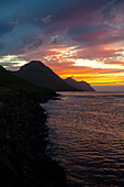 View of a dramatic sunset paints the sky with fiery hues over the dark, rugged coastline and tranquil waters, Syðradalur, Kalsoy, Faroe Islands.