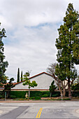 Blick auf eine ruhige Straße in der Nachbarschaft, in der üppige grüne Bäume ein cremefarbenes Gebäude unter einem wolkenverhangenen Himmel einrahmen und eine ruhige städtische Szene schaffen, Los Angeles, Kalifornien, Vereinigte Staaten.