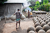 Munshiganj, Bangladesch - 23. November 2024: Blick auf einen Töpfer, der große Tontöpfe inmitten von Reihen von Töpferwaren trägt, während Kinder im dunstigen, sonnendurchfluteten Hof zusehen.