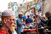 Tongi, Bangladesh - 31 January 2025: View of a bustling street scene, where vibrant textiles spill from a vendor's cart amidst a crowd of men in traditional caps near a colorful election billboard.