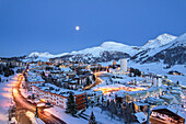 Aerial view of the softly illuminated town nestled among snow-laden peaks, with lights twinkling against the blue twilight sky, Sestriere, Piedmont, Italy.