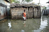 Kurigram, Bangladesh - 14 July 2024: View of a young boy wading through floodwaters near a simple home, the water reflecting the overcast sky and the textures of the surrounding dwellings.