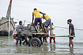 Cox's Bazar, Bangladesh - 01 January 2000: View of fishermen struggle with nets and a wooden cart through shallow waters to a vintage fishing boat.