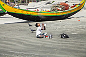 Cox's Bazar, Bangladesh - 08 February 2024: View of a boy drinking water near fishing boats on a sandy beach, flanked by crows and scattered belongings.