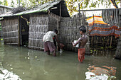 Kurigram, Bangladesch - 14. Juli 2024: Blick auf überschwemmte Häuser, in denen zwei Kinder im trüben Wasser stehen, deren Widerstandskraft in starkem Kontrast zu den baufälligen Gebäuden steht.