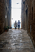 Pienza, Tuscany, Italy - 22 April 2025: View of a charming, narrow cobblestone street framed by aged stone buildings, leading to a bright horizon where a couple strolls hand-in-hand, birds soaring overhead.