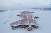 Aerial view of the snow-dusted island rises sharply from the frozen expanse, contrasting the stark white with its rugged, dark cliffs, Lake Baikal, Russia.