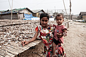 Cox's Bazar, Bangladesh - 07 February 2024: View of a girl holding a child amidst the stark reality of a refugee camp, where rows of drying fish contrast with the makeshift shelters.