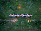 Aerial view of a long table set for a feast, nestled among lush green lawns and trees, illuminated by soft, warm lights, Assisi, Umbria, Italy.
