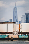 Blick auf hoch gestapelte Frachtcontainer mit dem One World Trade Center, das die Skyline unter einem wolkenverhangenen Himmel durchdringt, ein Tanz der Industrie und des Ehrgeizes, New York, New York, Vereinigte Staaten.