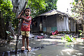 Noakhali, Bangladesh - 07 September 2024: View of a child standing on a log in floodwaters near a corrugated iron home, reflecting resilience amidst the challenging environment.