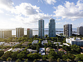 Luftaufnahme von glänzenden Wolkenkratzern, die die Skyline durchbrechen und im Kontrast zu den üppigen grünen Baumkronen darunter stehen, während in der Ferne die Biscayne Bay schimmert, Miami, Florida, Vereinigte Staaten.