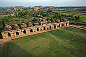 Aerial view of Elephant Stables, a long structure with domed roofs against a backdrop of verdant fields and rocky outcrops, Hampi, Karnataka, India.