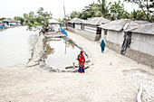Shyamnagar, Bangladesch - 13. Mai 2023: Blick auf eine Dorfszene, in der eine Frau in leuchtendem Rot und Blau an einem trüben Teich steht, der im Kontrast zu den gedämpften Tönen der Wellblechhäuser steht.
