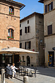 Pienza, Tuscany, Italy - 23 April 2025: View of quaint buildings with weathered facades, terracotta rooftops, and a sun-drenched cafe where patrons relax under a beige umbrella.