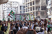 Chattogram, Bangladesh - 16 September 2024: View of jubilant crowds raising their hands towards a vibrant green bus adorned with flags and decorations, set against a backdrop of buildings.