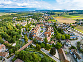 Luftaufnahme der Altstadt mit ihren roten Ziegeldächern und der Holzbrücke über die Aare, eingebettet in grüne Bäume und goldene Felder, Aarberg, Kanton Bern, Schweiz.