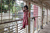 Noakhali, Bangladesh - 02 September 2024: View of a young girl playfully hangs from a window, floodwaters reflecting the overcast sky, creating a somber scene.