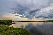 Blick auf einen ruhigen Fluss, in dem sich der dramatische, wolkenverhangene Himmel spiegelt, umrahmt von üppiger grüner Vegetation, die eine ruhige und doch kraftvolle Szene schafft, Masindi, Nordregion, Uganda.