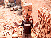 Narsingdi, Bangladesh - 08 December 2024: View of a man balancing bricks on his head amidst the terracotta tones of a brickyard, contrasting with the pale sky.