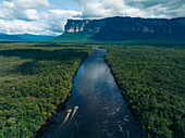 Aerial view of a dark Churún river snaking through a dense emerald jungle towards the towering tepuis, their flat tops kissed by the clouds, Canaima, Bolívar, Venezuela.