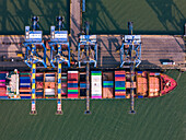 Luftaufnahme eines Containerschiffs, das im Hafen angedockt ist. Unter den hoch aufragenden blauen Kränen stapeln sich pulsierende Container, die industrielle Aktivität im Jawaharlal Nehru Port Trust (JNPT) in Navi Mumbai, Indien, ausstrahlen.