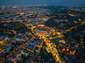 Luftaufnahme der antiken Steine des Kolosseums, die in der kühlen blauen Dämmerung warm leuchten, umgeben von der beleuchteten Stadtlandschaft, Rom, Latium, Italien.