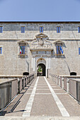 View of a long stone bridge leading to a grand entrance flanked by Italian and EU flags on a light stone facade under a clear sky, L'Aquila, Abruzzo, Italy.